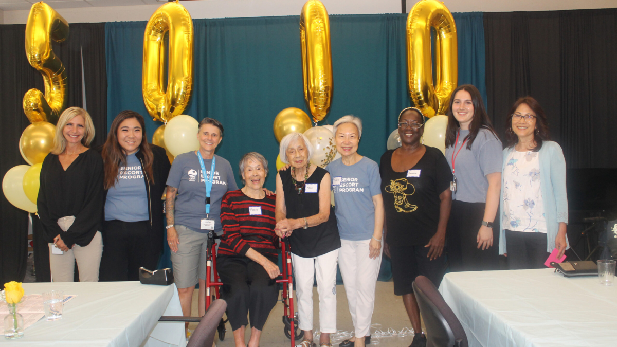 A group of nine people stand smiling in front of large gold 30000 balloons, celebrating at an indoor event with decorated tables and balloons.
