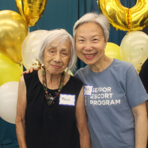 Two older women smile together at an event, standing in front of gold and white balloons. One wears a black top and necklace, the other a blue Senior Escort Program shirt. Both have nametags.