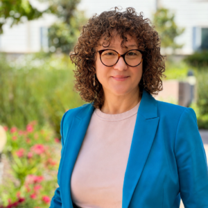 A person with curly hair and glasses, wearing a blue blazer and light shirt, stands outside in a garden with greenery and flowers, smiling at the camera.