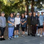 A group of thirteen adults stands smiling outdoors under tall trees during the Ohana Walk fundraiser, dressed in casual sporty clothes and hats, with sunlight filtering through the leaves as they support ACC Senior Services.