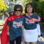 Two smiling women wearing Ohana Walk shirts walk outside during the Ohana Walk fundraiser for ACC Senior Services; one wears a red cape, the other a white skirt and a colorful lei. Other participants stroll behind them on a sunny path surrounded by trees.
