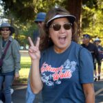 Smiling woman wearing sunglasses and a visor flashes a peace sign while walking outdoors at an Ohana Walk fundraiser for ACC Senior Services. Others in similar shirts walk behind her in a sunny, tree-filled park.