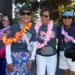 Four women in sunglasses and leis smile outdoors at a park event during the Ohana Walk fundraiser for ACC Senior Services. They wear matching shirts and crossbody bags, standing on a sunny path with trees in the background.