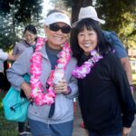 Two smiling women wearing sunglasses and colorful flower leis pose together outdoors at the Ohana Walk fundraiser for ACC Senior Services. Others are in the background, and tall trees surround the group on a sunny day.