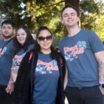Four people wearing matching Dhama Walk shirts stand together outdoors, smiling at the camera, with trees and sunlight in the background during the Ohana Walk fundraiser for ACC Senior Services.