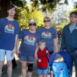 Five people pose outdoors near trees and sunlight at the Ohana Walk fundraiser supporting ACC Senior Services. Three adults wear matching “Ohana means family” shirts, a child in a Spider-Man shirt stands smiling, and a stroller with a Stitch design is in front.
