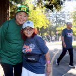 Two smiling people pose arm-in-arm on a park path during the Ohana Walk fundraiser for ACC Senior Services. Others walk behind them wearing event T-shirts, while trees and sunlight fill the background.