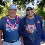 Two older men smiling and walking outdoors at a park during the Ohana Walk fundraiser, both wearing matching event shirts supporting ACC Senior Services. One wears a flower lei, while the other sports sunglasses and a hat. Trees and other participants are visible in the background.