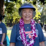 Older woman wearing a blue visor, purple flower lei, and blue T-shirt smiles outdoors at the Ohana Walk fundraiser for ACC Senior Services. Other people, also in blue shirts, walk nearby under trees in the background.