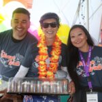 Three people wearing Ohana Walk shirts smile at a festive event in support of the Ohana Walk fundraiser for ACC Senior Services. One holds a cake, another wears a flower lei and sunglasses, and the third has a name badge. Colorful balloons are in the background.