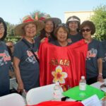 A group of smiling women in matching shirts pose outdoors at the Ohana Walk fundraiser, with one holding a red cloth that says KAKATA and features a large flower design. Water bottles and papers from ACC Senior Services sit on the table in front of them.