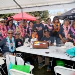 A group of people wearing leis and matching shirts gather around a table with a decorated cake at an outdoor event under a tent, celebrating the Ohana Walk fundraiser for ACC Senior Services.