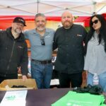 Four adults stand together under tents at the Ohana Walk fundraiser for ACC Senior Services, smiling at the camera behind a table with green bags and a cardboard box. Banners and people are visible in the background.