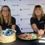 Two women in black shirts sit at a table with baskets of photos and colorful treats, volunteering for the Ohana Walk fundraiser benefiting ACC Senior Services. A banner behind them shows Compass and Catricala Real Estate Group logos. Both women are smiling at the camera.
