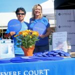 Two smiling people in blue shirts stand behind a promotional table with frisbees, a flower pot, and pamphlets, proudly supporting the Ohana Walk fundraiser. The tablecloth reads Revere Court, representing ACC Senior Services. Event booths and banners are visible in the background.