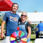 Two people stand smiling at an outdoor event beside a colorful prize wheel, under a red canopy at the Ohana Walk fundraiser for ACC Senior Services. White tents, tables, and attendees are visible in the sunny background.