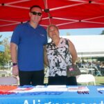 A man and woman smile at an outdoor event booth under a red tent during the Ohana Walk fundraiser for ACC Senior Services. The table is set with brochures, pens, and a water bottle, while people sit at tables in the background.