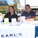Two people sit at an outdoor event booth for Pearls Nutrition Management Services, smiling at the camera during the Ohana Walk fundraiser hosted by ACC Senior Services. The table displays marketing materials and a plant, while other attendees are seated at tables in the background.