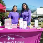 Two women in purple shirts stand behind a table with informational materials and donation boxes for My Sisters House at the Ohana Walk fundraiser organized by ACC Senior Services, held at an outdoor event with tents and people in the background.