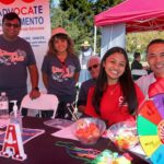 Five people smile at an outdoor event booth with a colorful prize wheel, brochures, candy, and hand sanitizer on the table. A banner behind them reads Advocate Sacramento. This cheerful gathering takes place at the Ohana Walk fundraiser for ACC Senior Services, with trees and tents visible in the background.