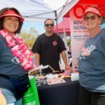 Three people smile at an outdoor event booth during the Ohana Walk fundraiser for ACC Senior Services. Two women, wearing leis and visors, stand in front of a table covered with flyers and pins. A man stands behind the table under a tent decorated with a banner. The atmosphere is sunny and festive.