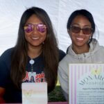 Two women wearing sunglasses sit at a table with Monarch Hospice signs in front of them at the Ohana Walk fundraiser for ACC Senior Services. They are smiling at the camera, and a white tent background is visible behind them.