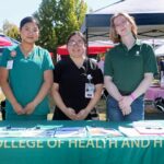 Three people stand behind a table with clipboards, outdoors under tents during the Ohana Walk fundraiser. The tablecloth reads COLLEGE OF HEALTH AND HUMAN. Two wear scrubs with badges, and one wears a casual shirt, representing ACC Senior Services. All are smiling at the camera.