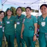 Five smiling people in green medical scrubs stand together outdoors under a white canopy, some with stethoscopes and name badges, posing for a group photo at the Ohana Walk fundraiser for ACC Senior Services.