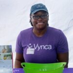 A person wearing a Vynca shirt and hat smiles at a booth with informational flyers and a green container, supporting the Ohana Walk fundraiser for ACC Senior Services. They have a name tag that says Christine.