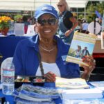 Smiling woman in a blue shirt and hat sits at a table with brochures and a meal, holding up a California State Retirees booklet at an outdoor event for the Ohana Walk fundraiser hosted by ACC Senior Services.