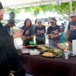 A chef in black attire demonstrates cooking techniques to a group of people gathered around a table with food items under an outdoor tent at the Ohana Walk fundraiser for ACC Senior Services. The audience, all attentively watching and wearing matching event t-shirts, enjoys the culinary demonstration as part of this special community event.