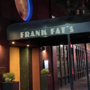 Restaurant entrance at night with a black awning displaying FRANK FATS in white letters. The entrance is flanked by two potted plants and there is a dimly lit circular sign above.
