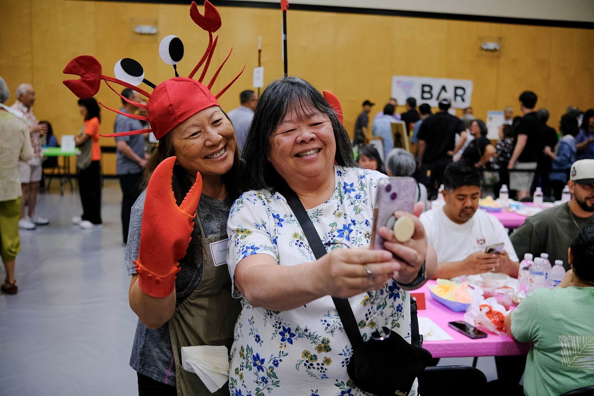 Two smiling women take a selfie, with one wearing a red crab hat and claw mittens at a community event.