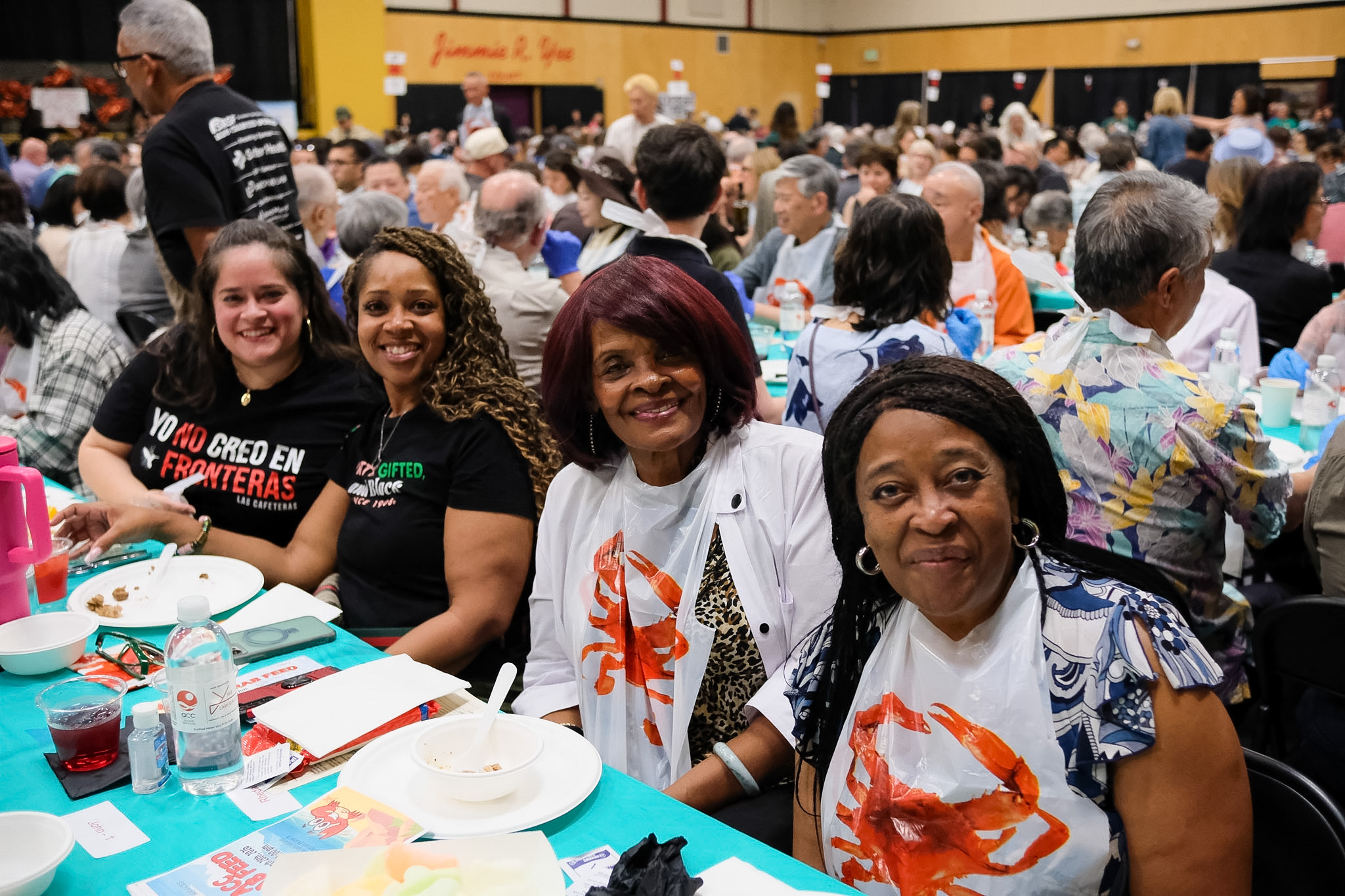 Four smiling women sit at a table covered with a teal cloth at a crowded banquet, wearing white bibs decorated with an orange crab graphic.