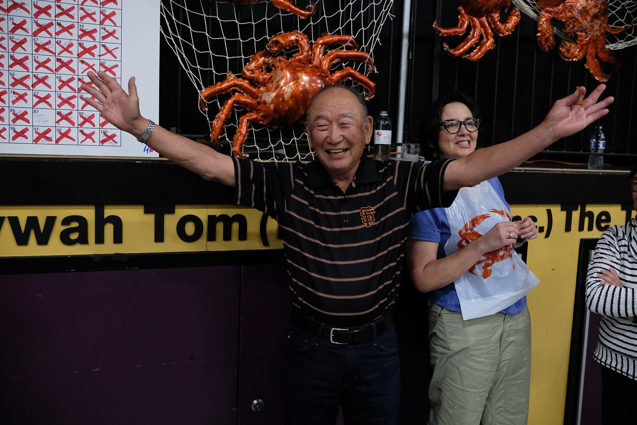 An elderly man with arms outstretched smiling next to a woman, in front of a raffle board and a wall decoration of a large plastic crab.