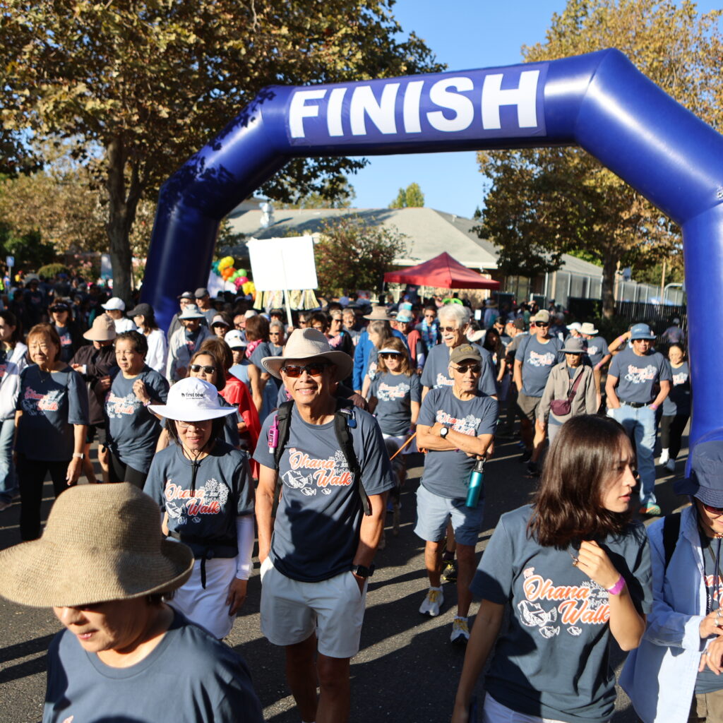 A crowd of people wearing matching navy blue Ohana Walk t-shirts walk under a large blue inflatable finish line arch at an outdoor event.