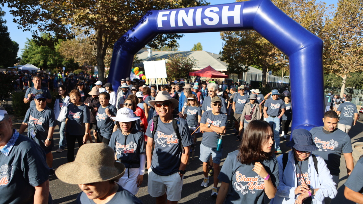 A crowd of people wearing matching navy blue Ohana Walk t-shirts walk under a large blue inflatable finish line arch at an outdoor event.