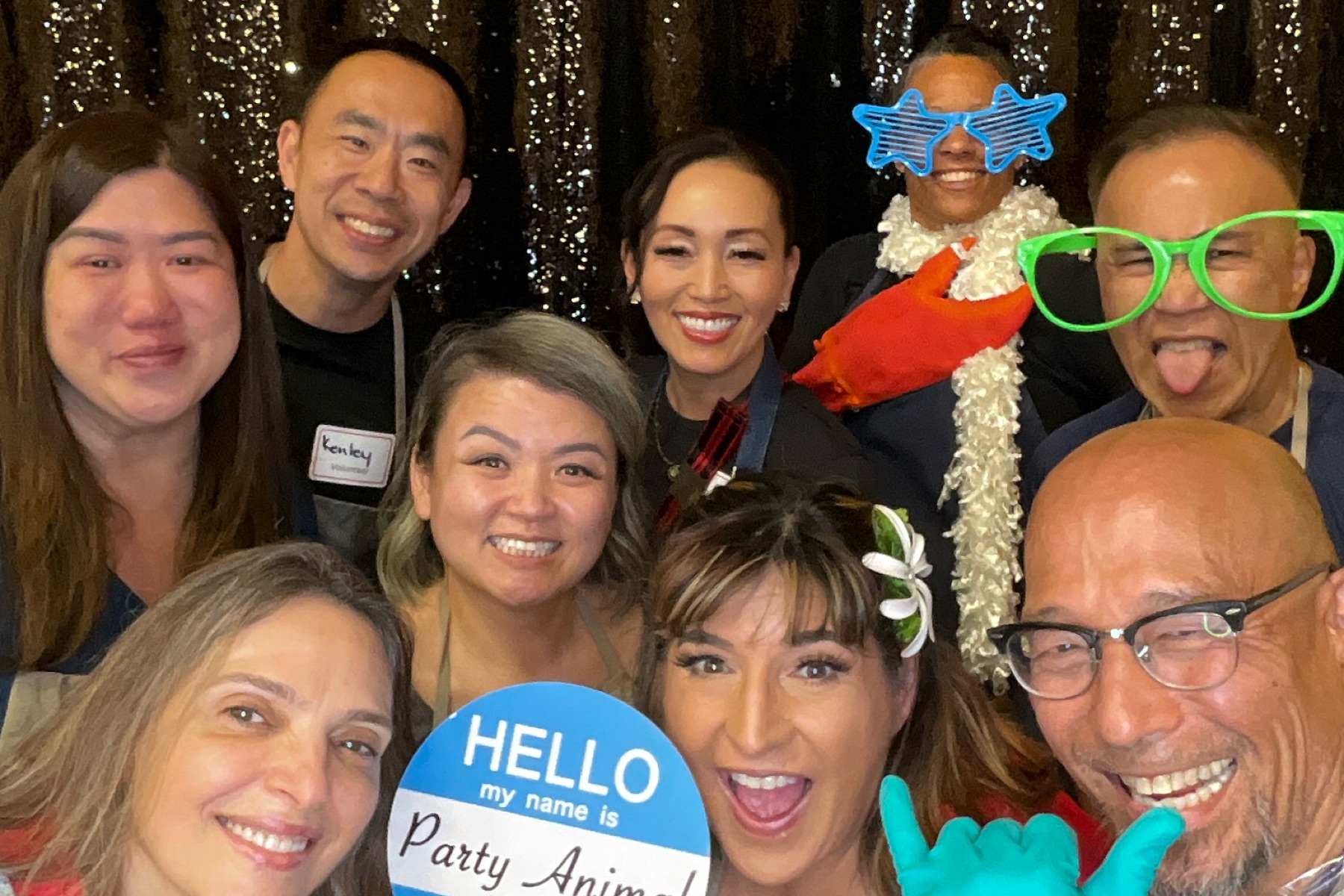 A group of people posing for a photo, some wearing party props including oversized glasses, a flower hair clip, and a foam lobster claw.