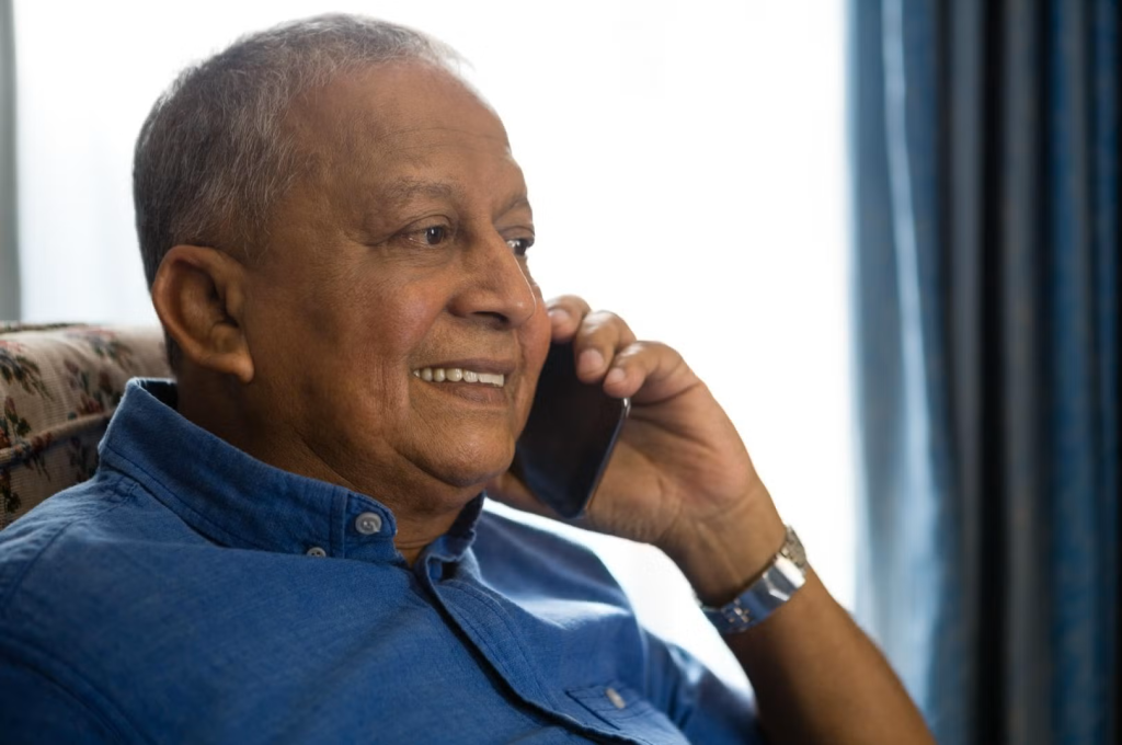 An elderly man with short gray hair, wearing a blue collared shirt, sits indoors and smiles while talking on a smartphone. He is seated on a patterned chair near a window with blue curtains.